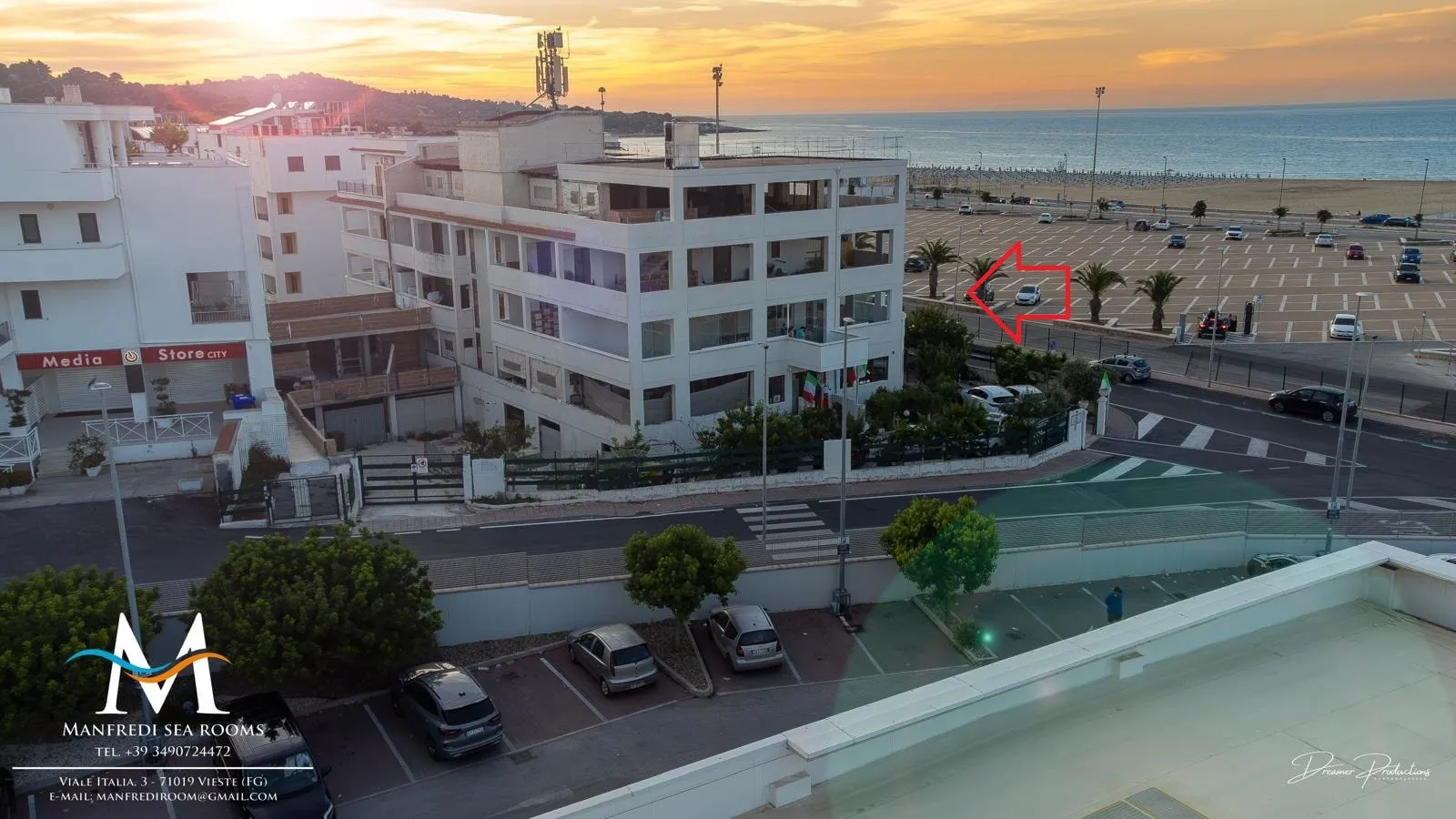 Veduta aerea al tramonto: l’edificio con le verande, il piazzale con le palme, la spiaggia e il mare. Una freccia rossa indica il piazzale