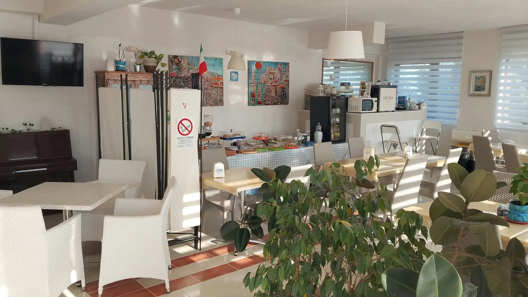 La sala colazione: tavoli, pianoforte, piante e il buffet in fondo